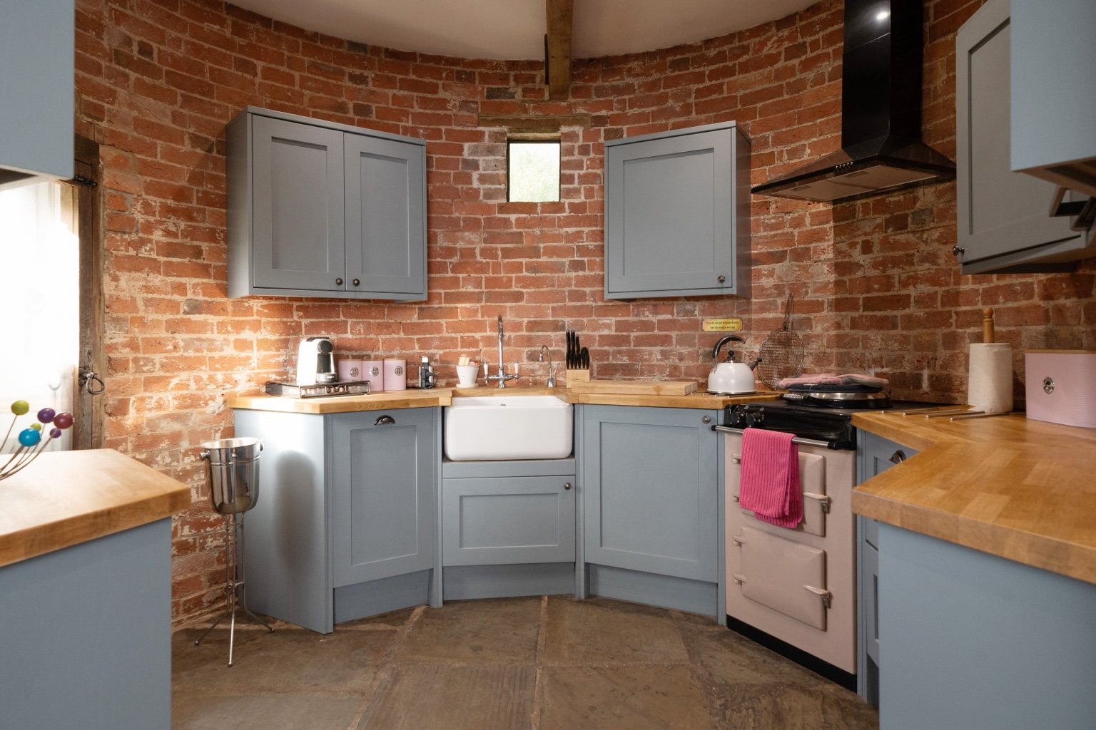 The round kitchen at The Oast, with exposed brick walls, blue-grey cabinets, a Belfast sink and a cream AGA with black lids